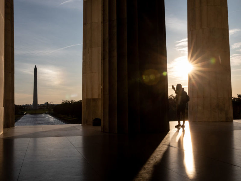 a woman is backlit by the sun as she takes a photo of the washington monument from the lincoln memorial