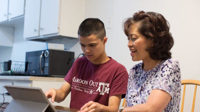 a mother and son, who is wearing a maroon out t-shirt, sitting at a table looking at a tablet