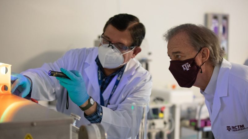 a scientist in a lab points to something to show john sharp, both wearing masks