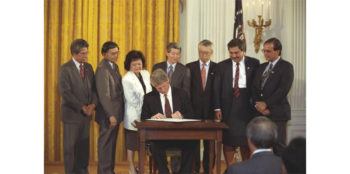 bill clinton seated at a desk surrounded by people as he signs a document