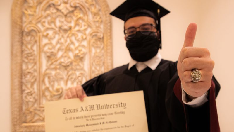 student wearing cap and gown holds his diploma in one hand and shows his aggie ring with the other giving a gig 'em