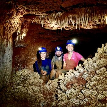 two students and professor wearing headlamps stand in a cave surrounded by stalactites