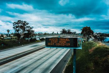 a photo of an empty highway with a message on a traffic sign that says Avoid Travel, Stay At Home, Beat COVID-19