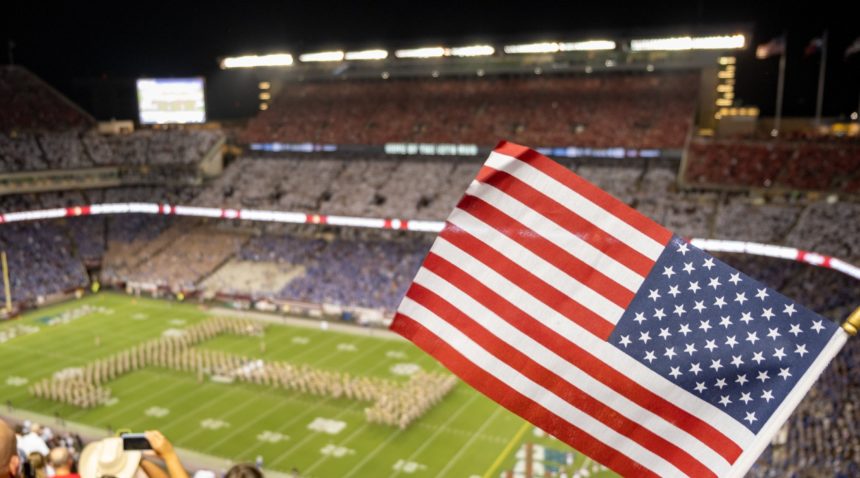 an american flag flies in front of a scene overlooking kyle field, where fans where red white and blue shirts