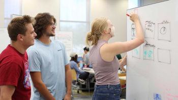 two male students watch as a female student writes ideas on a white board