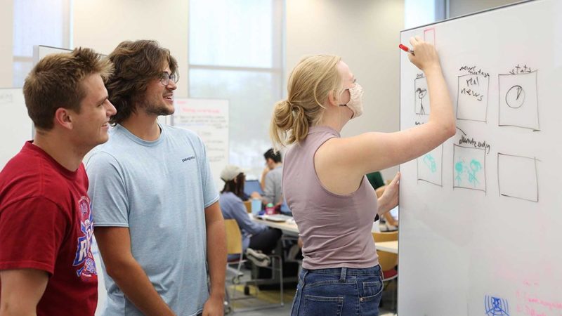 two male students watch as a female student writes ideas on a white board