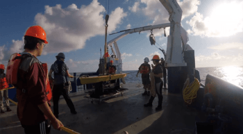 video still of a group of students in hard hats on a boat preparing to launch a yellow glider into the water