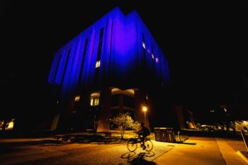 The Harrington Education Center Office Tower lit with blue lights as a person on a bicycle cycles past at night