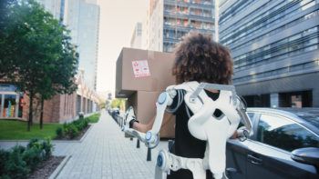 woman wears a lower back exoskeleton device while carrying a moving box down a sidewalk