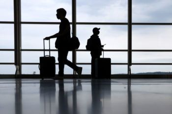silhouette of two travelers wearing face masks pushing their suitcases through an airport