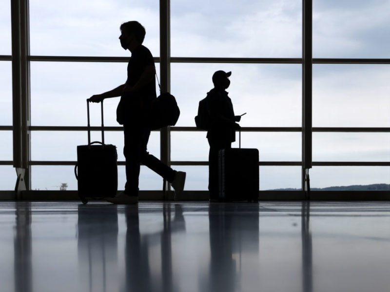silhouette of two travelers wearing face masks pushing their suitcases through an airport
