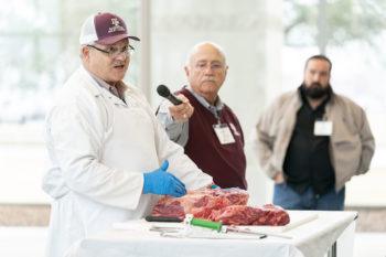 a man wearing a white coat and gloves gives a barbecue demonstration using a cut of beef sitting on a table before him