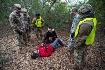 students in a wooded area stand over a man lying on the ground giving emergency medical treatment