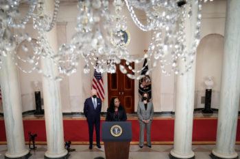 view through a chandelier in a room where the president and vice president stand behind jackson at a podium as she makes remarks