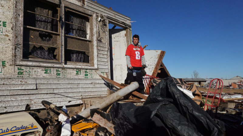 a man stands on the porch in front of a house destroyed by a tornado