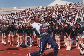 man wearing overalls leading a yell in front of a crowd of texas a&m students on the sidelines