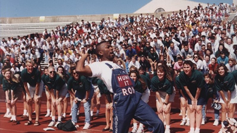 man wearing overalls leading a yell in front of a crowd of texas a&m students on the sidelines