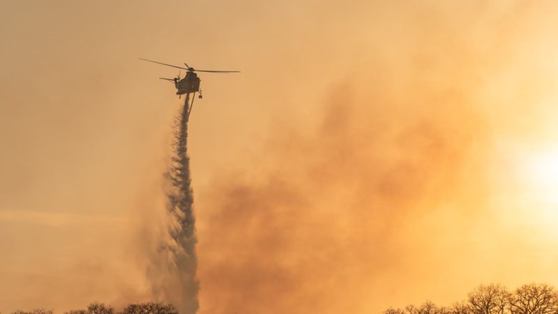 silhouette of a helicopter dumpin gwater over a wildfire against an orange sunset