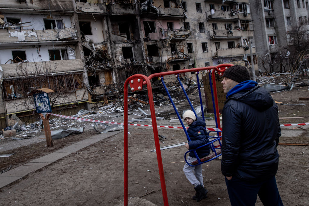a young boy sits on a swing in front of a severely damaged residential building