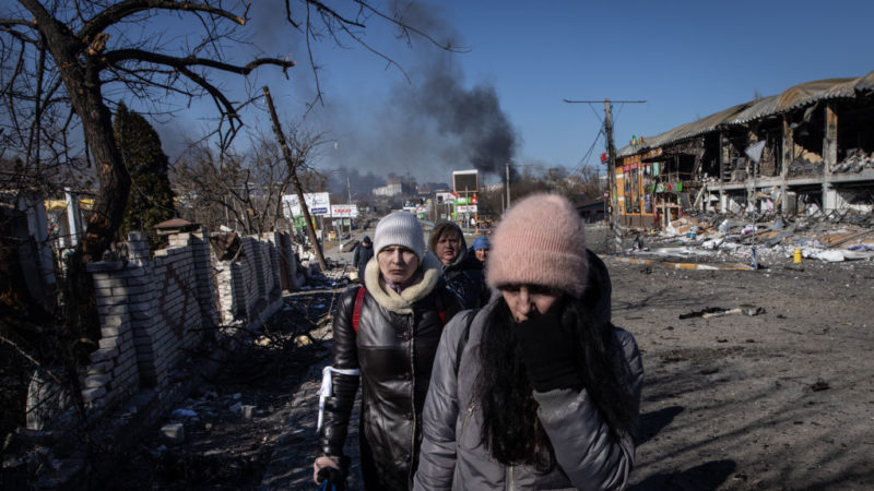 a group of civilians walk away from a city with smoke rising in the background among destroyed buildings