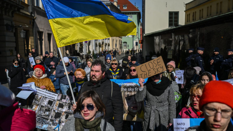 a crowd of people stand in a street protesting in favor of a no-fly zone. some are holding signs, and a ukrainian flag