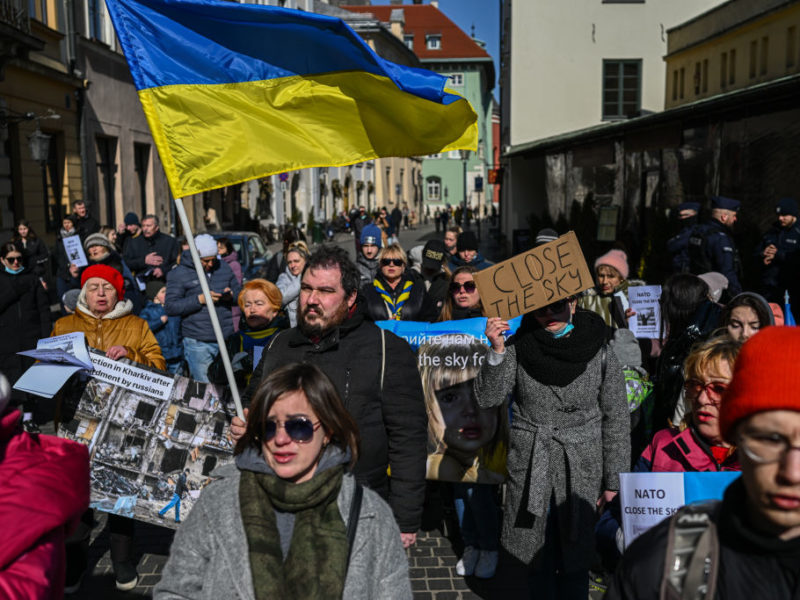 a crowd of people stand in a street protesting in favor of a no-fly zone. some are holding signs, and a ukrainian flag