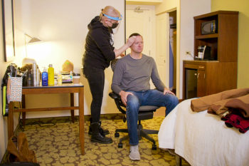 Laurent Therivel sits down in his hotel room as a makeup artist begins to apply his disguise