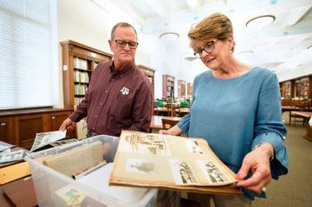 a woman leafs through a scrapbook containing old photographs while a man next to her looks on