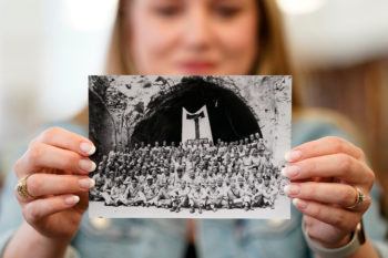 a woman holds up a black and white photograph of a group of uniformed men posing in front of a tunnel, where a large flag that says "aggies" hangs