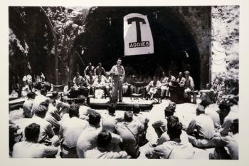 a black-and-white photo showing a group of uniformed men gathered at the moth of a tunnel watching a man in the center speak from a podium