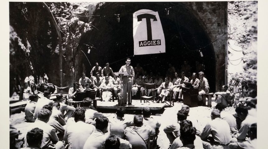 a black-and-white photo showing a group of uniformed men gathered at the moth of a tunnel watching a man in the center speak from a podium