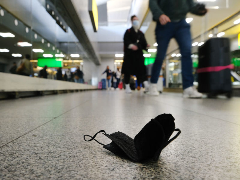 a photo of a black face mask sitting on the floor in the foreground as people with suitcases walk by in the background