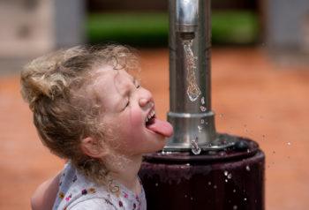 a little girl stands under a spigot of running water holding her tongue out