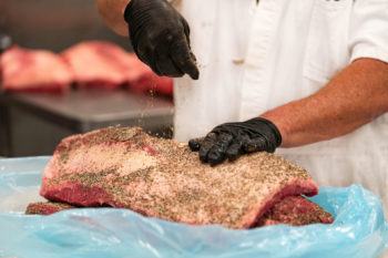 cropped image of a man wearing gloves standing in front of a piece of meat as he applies seasoning