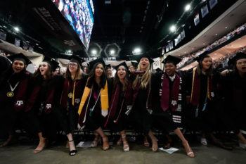 a group of students in graduation regalia link arms at commencement in reed arena