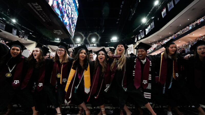 a group of students in graduation regalia link arms at commencement in reed arena