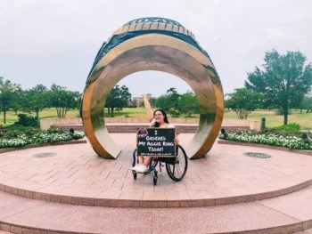 a picture of a woman in front of a large model of an Aggie Ring holding an "I ordered my Aggie Ring today!" sign