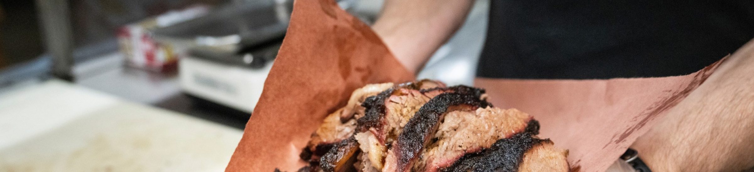 close up image of a man holding out a brown piece of butcher paper piled with slices of brisket