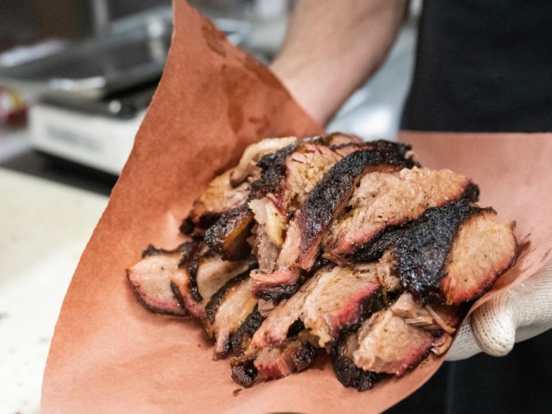 close up image of a man holding out a brown piece of butcher paper piled with slices of brisket