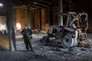 a photo of a man in olive green clothes standing with his hands on his hips in the middle of a large warehouse full of holes and debris. to the right of him is the burned out husk of a tractor. to the left is a pile of corn scattered across the floor.