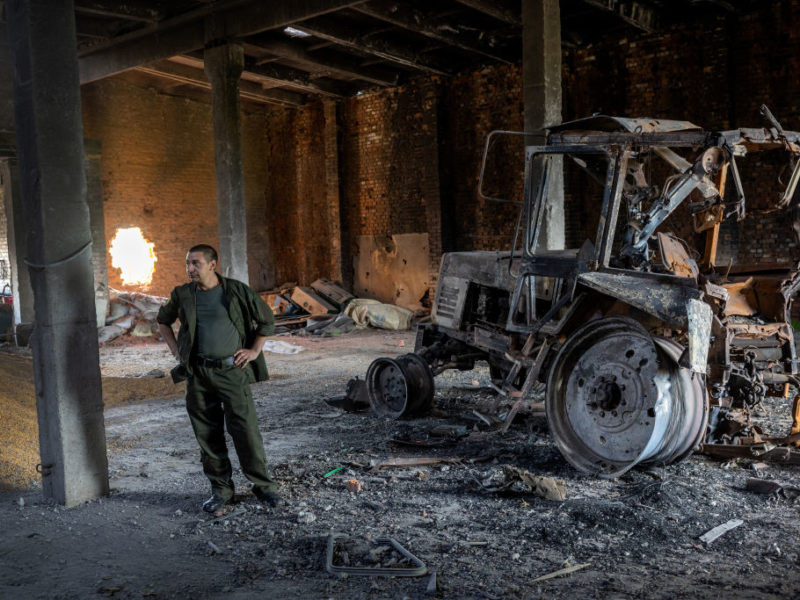 a photo of a man in olive green clothes standing with his hands on his hips in the middle of a large warehouse full of holes and debris. to the right of him is the burned out husk of a tractor. to the left is a pile of corn scattered across the floor.