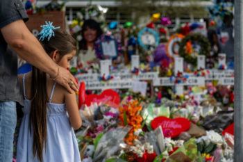 a photo of a girl in front of a large pile of flowers, wreaths, crosses and other items next to the sign for Robb Elementary School in Uvalde, Texas.  A man stands to her left with his hand on her shoulder.