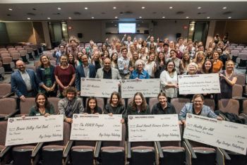 a group photo of students in a lecture hall giving the thumbs up behind a group of nonprofit representatives holding giant checks and smiling