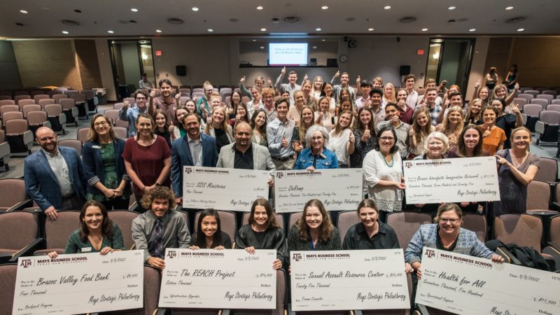 a group photo of students in a lecture hall giving the thumbs up behind a group of nonprofit representatives holding giant checks and smiling