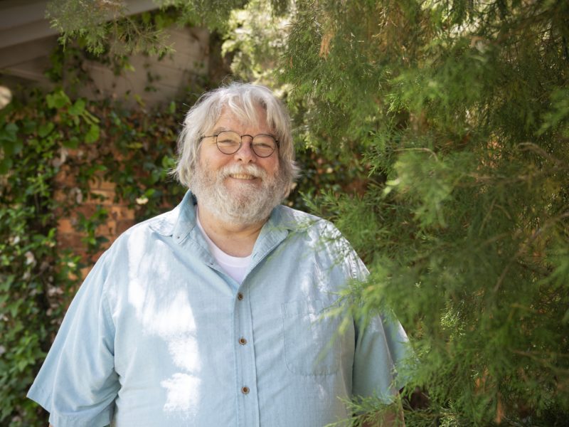 a photo of a man with glasses, white hair and a white beard smiling among some plants
