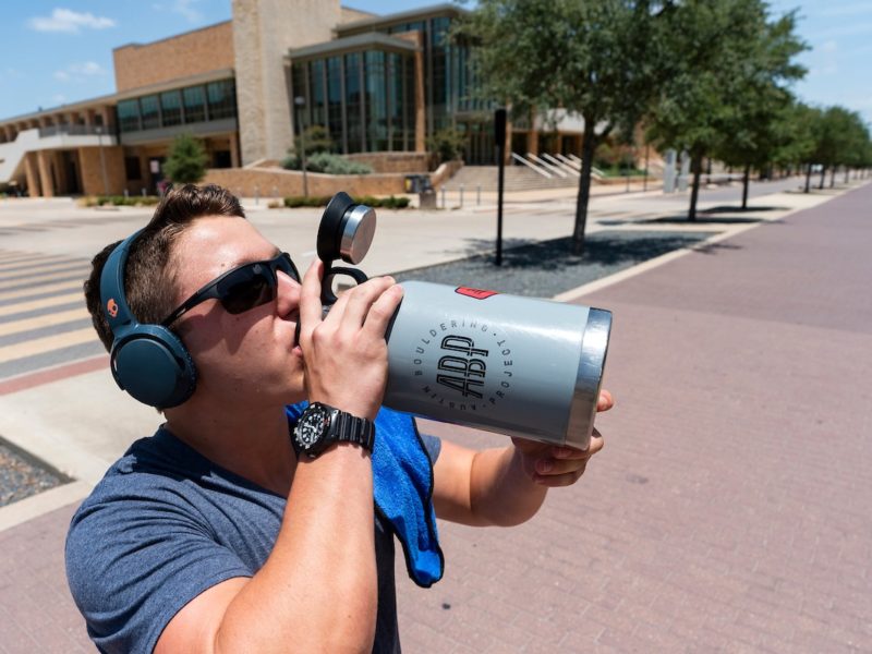 A man on campus wearing sunglasses takes a drink out of a large blue water bottle