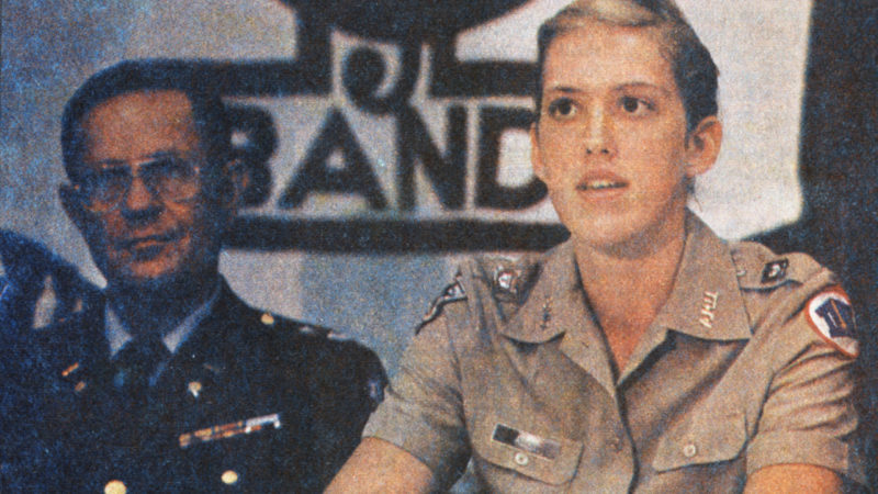 a photo of a young woman in a corps of cadets uniform sitting at a table with an older man wearing glasses and a dark military uniform. behind them is the bottom half of an Aggie Band banner