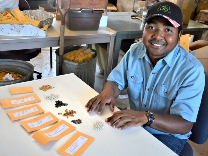 a photo of a man in casual clothes and a cap sitting at a table in front of various piles of different colored beans