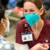 a female nursing student wearing maroon scrubs and a face mask kneels down in front of a patient