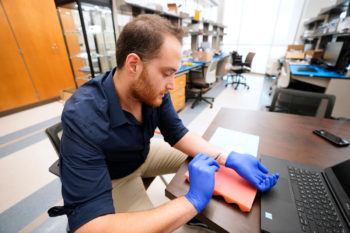 A man seated at a lab table wearing gloves uses forceps to apply a tiny strip of paper onto his wrist